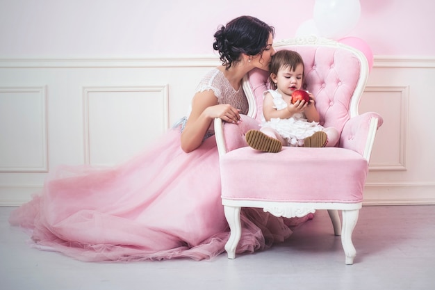 Mother and daughter a beautiful and happy pink interior with vintage chair and balls in beautiful dresses holiday