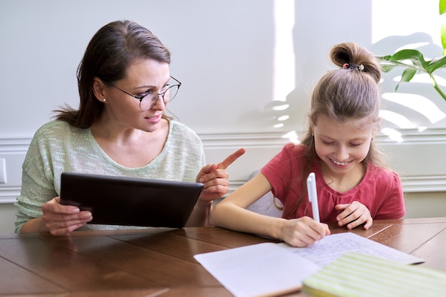 Mother and daughter child study together at home, sitting at table. Woman with digital tablet, using Internet, girl writing in notebook. Distance learning, parent helping child primary school student