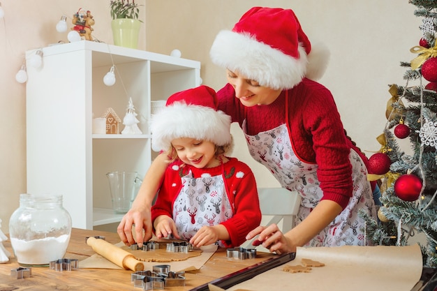 Mother and daughter cut out Christmas gingerbread cookies for happy holidays in Santa hats