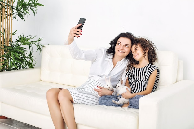Madre e figlia felici con soffici conigli seduti su un divano bianco e facendo selfie sul cellulare