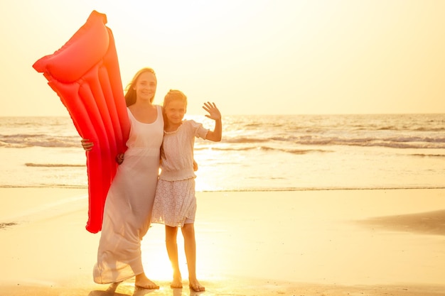 Mother and daughter having fun walking by summer sunset beach with air mattress in the sea clear blue water