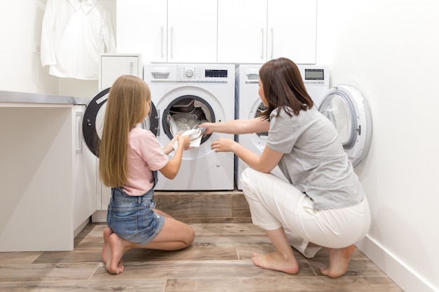 洗濯機と乾燥機の近くのランドリールームで母と娘のヘルパーがきれいな服を脱いで