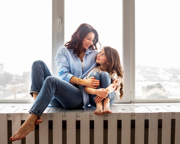 Photo mother and daughter hugging on window sill