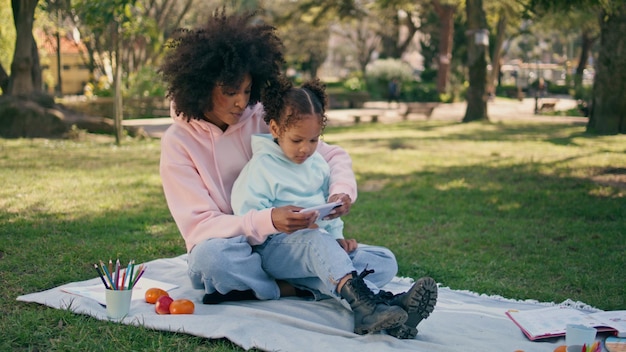 Photo mother daughter looking smartphone at picnic park mom showing cartoon to girl