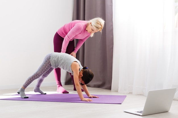 Mother and daughter practicing online yoga lesson at home at quarantine isolation period during coronavirus pandemic. Family doing sport together online from home. Healthy lifestyle