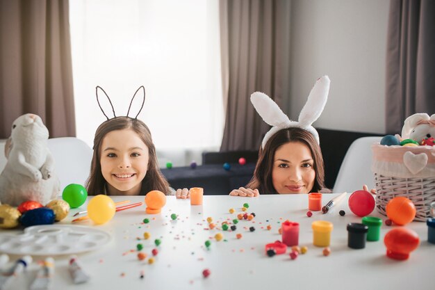 Mother and daughter prepare for Easter. They hide behind table with decoration, eggs and painting on it. Mother and daughter look straight and smile. They wear bunny ears.