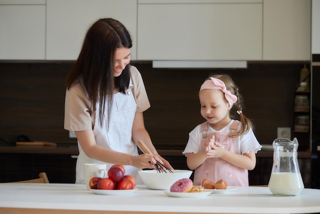 Mother and daughter preparing a sweet cake using flour milk sitting on chairs at a table in a modern