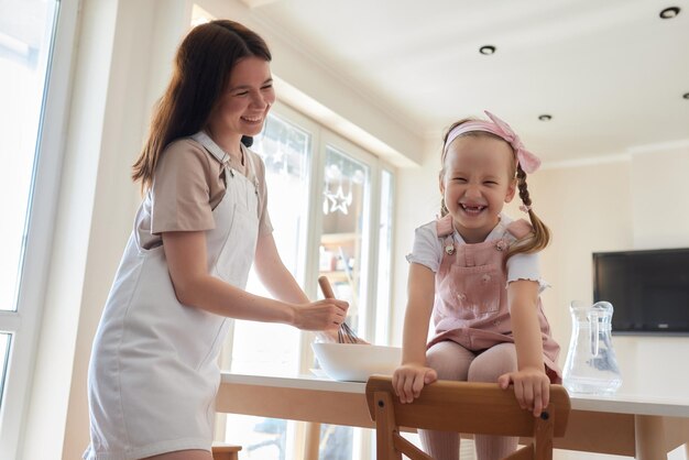 Mother and daughter preparing a sweet cake using flour milk sitting on chairs at a table in a modern