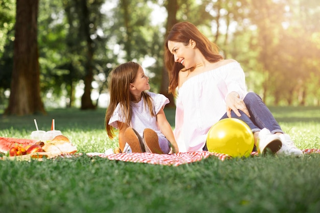 Mother and daughter sitting on the blanket in the park and looking at each other