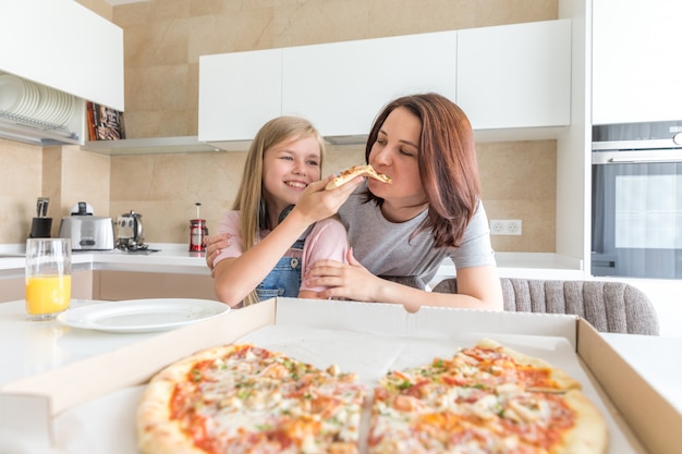 Mother and daughter sitting in the kitchen, eating pizza and having fun