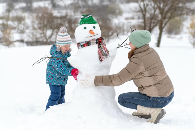 Mother and daughter or son playing with snowman outside Cute child with mon making snowman in the park