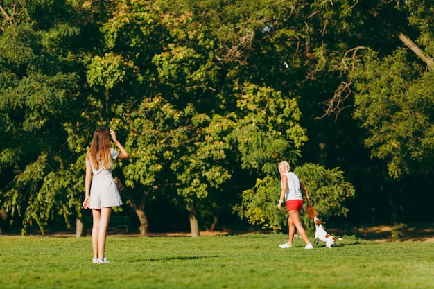 Mother and daughter throwing orange flying disk to small funny dog, which catching it on green grass. Little Jack Russel Terrier pet playing outdoors in park. Dog and women. Family resting on open air