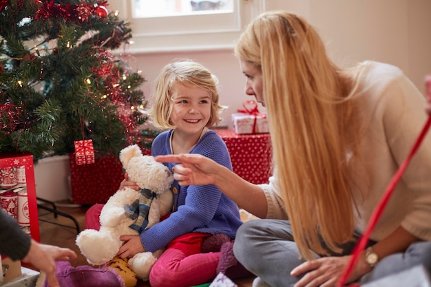 テディベアを持ったクリスマス ツリーの下でプレゼントの包装を解く母と娘