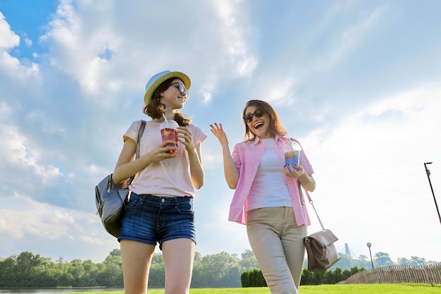 公園の芝生の上を歩く母と娘、親と子のティーンエイジャーの関係、晴れた夏の日。飲み物、雲の背景の青い空と幸せな女性