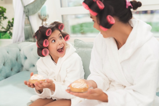 Mother and daughter in white bathrobes are sitting on couch.