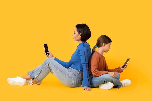 Mother And Daughter With Smartphones Sitting Back To Back On Yellow Background