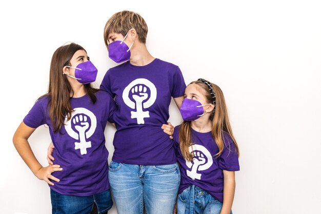 Mother and daughters on a white wall and wearing a purple T-shirt with the symbol of working women on International Women's Day, March 8, with a face mask for the coronavirus pandemic