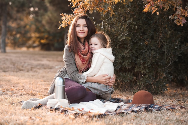 Mother drinking tea with kid daughter  sitting in sun light in park with picnic