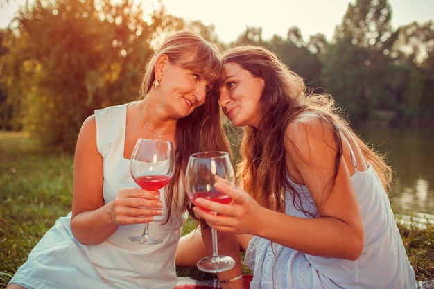 Mother drinking wine with her adult daughter by summer river at sunset. 