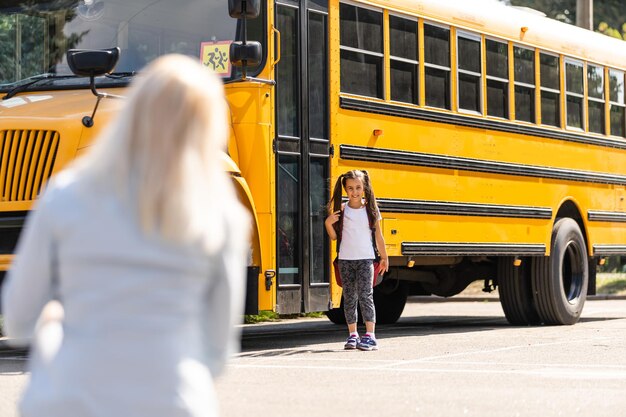 Mother escorts the schoolgirl with ponytails and a backpack to school.