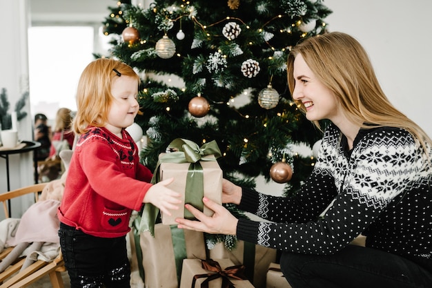 母は娘と贈り物を交換します親と子はクリスマスツリーの近くで楽しんでいます