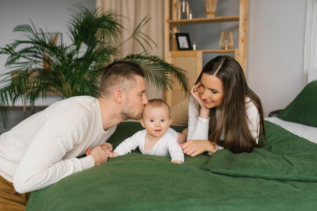 Mother and father are lying on the bed with their little son at home Dad kisses his baby