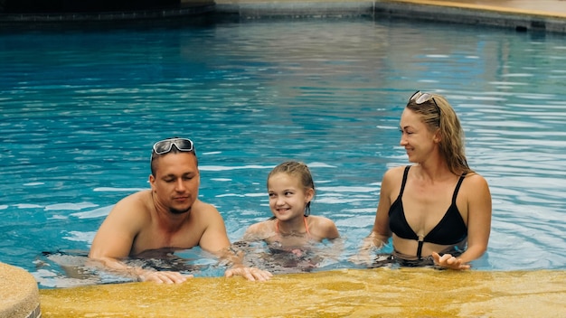 The mother and father with little daughter have fun in the pool. Mom and dad plays with the child. The family enjoy summer vacation in a swimming pool jumping, spinning, splash water. Slow motion.