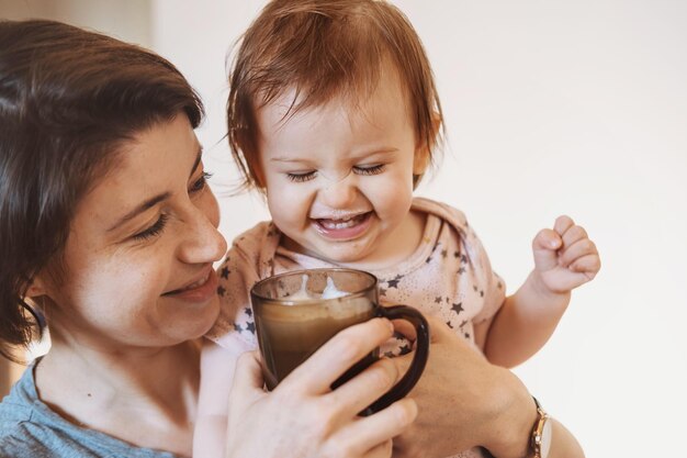 Madre che alimenta la bambina con una tazza di latte in piedi a casa concetto di maternità sorridente bambino felice