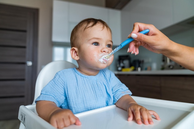 Mother feeding baby with spoon in kitchen Baby eating Portrait of happy young baby boy in high chair