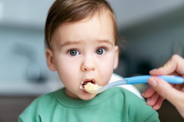 Mother feeding baby with spoon in kitchen Small child sits on a chair and eating