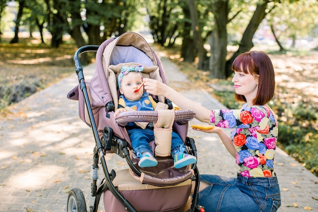 スプーンで彼女の女の赤ちゃんを授乳する母親。彼女の愛らしい子供に食べ物を与える母。かわいい赤ちゃんのベビーカーキャリッジの上に座って、笑顔のポーズ