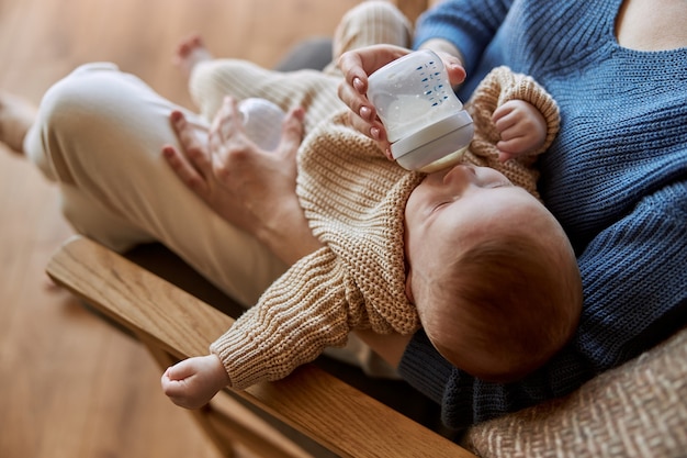 Мать кормит своего новорожденного ребенка из бутылочки с молоком. Женщина и европейский младенческий ребенок, сидя в деревянном кресле. Интерьер однокомнатной квартиры. Понятие материнства и ухода за ребенком
