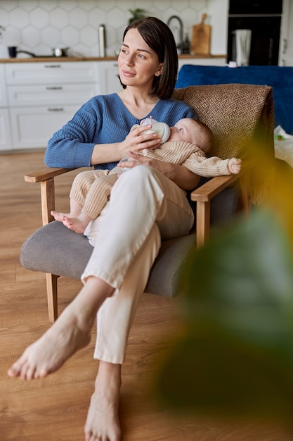 Mother feeding her newborn baby from bottle with milk. Young thoughtful european woman and infant kid sit in wooden armchair. Interior of studio apartment. Concept of motherhood and child care