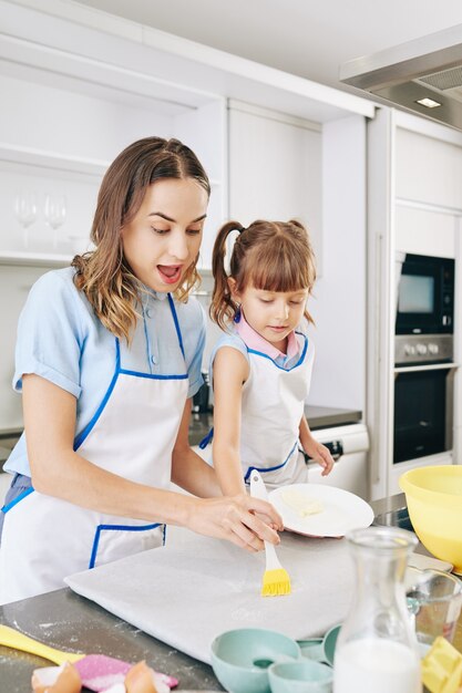 Mother helping daughter to apply melted butter on parchment paper with silicone brush