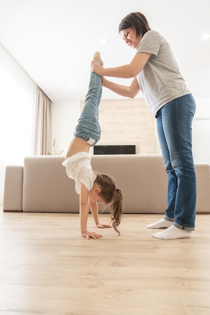 Mother helping daughter to walk upside down on her arms at home holding legs