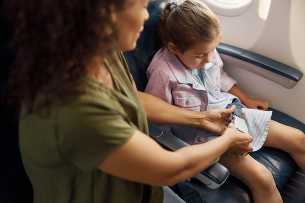 Madre che aiuta la sua piccola figlia a regolare e stringere la cintura di sicurezza su un aereo per un volo sicuro. viaggiare in aereo durante covid19, concetto di famiglia