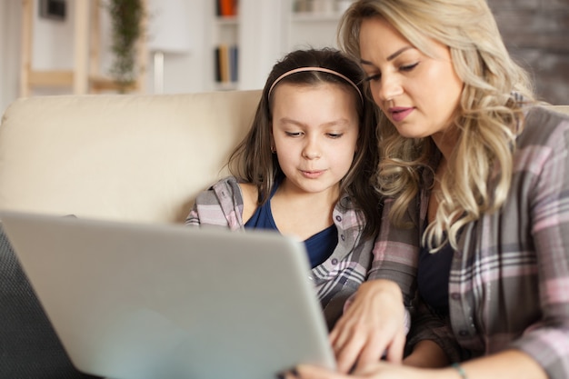 Mother helping little daughter to type on laptop keyboard sitting on the ouch in living room.