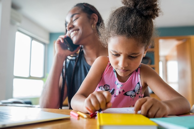 Mother helping and supporting her daughter with online school while talking on the phone at home. New normal lifestyle concept. Monoparental concept.