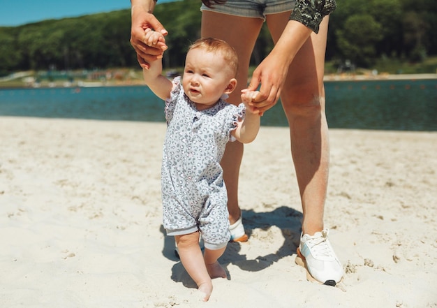 母は女の子がビーチで最初の一歩を踏み出すのを手伝います夏の日の前景で砂の上を一緒に素敵に歩く