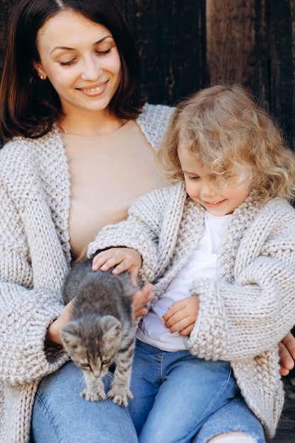 Mother and her daughter play with a little cat by the wooden wall.