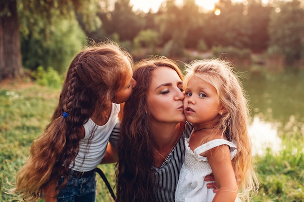 Mother and her daughters hugging and kissing outdoors. Family walking by summer river at sunset.