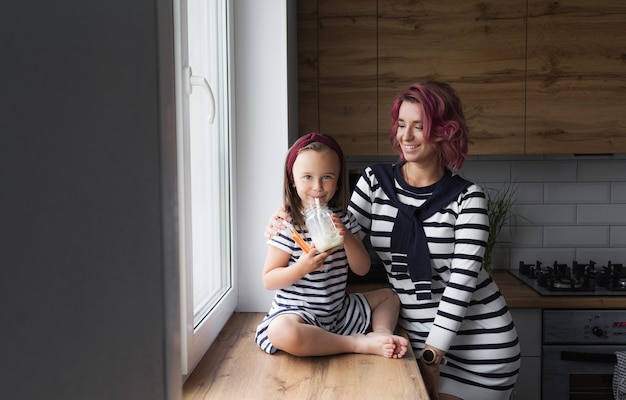 mother and her little daughter are sitting on the windowsill in the kitchen and drinking milk