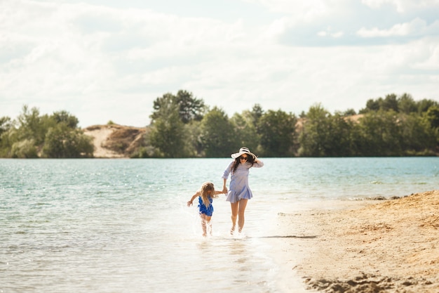 母と彼女の小さな娘が海岸で楽しんでいます。若い可愛いお母さんと彼女の子供が水の近くで遊んで