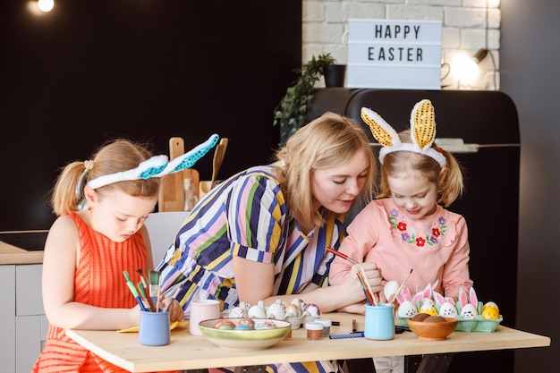 Foto la madre e le sue due figlie stanno decorando le uova di pasqua per la celebrazione delle vacanze di pasqua a casa nella loro cucina. congratulazioni di buona pasqua sul tablet sullo sfondo.