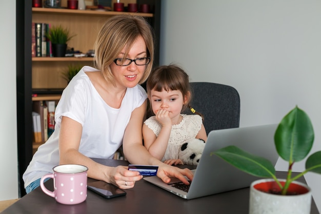 Фото Мать держит кредитную карту с маленькой дочерью, сидя рядом, глядя на ноутбук. концепция покупок в интернете.