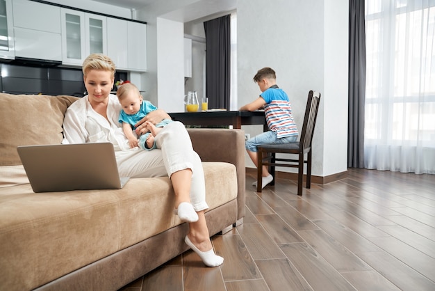 Mother holding baby while sitting on couch with laptop