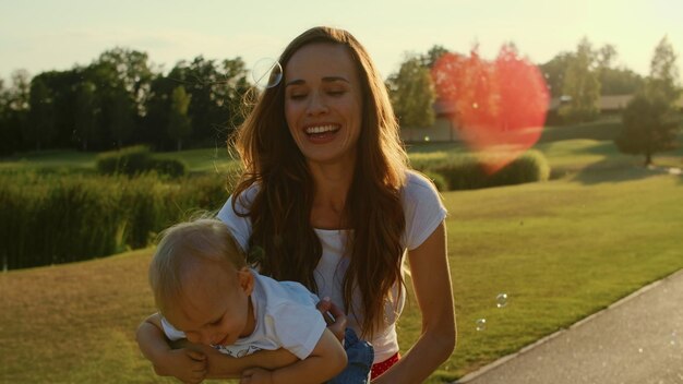 Photo mother holding little son on hands in parktoddler looking soap bubbles outdoors