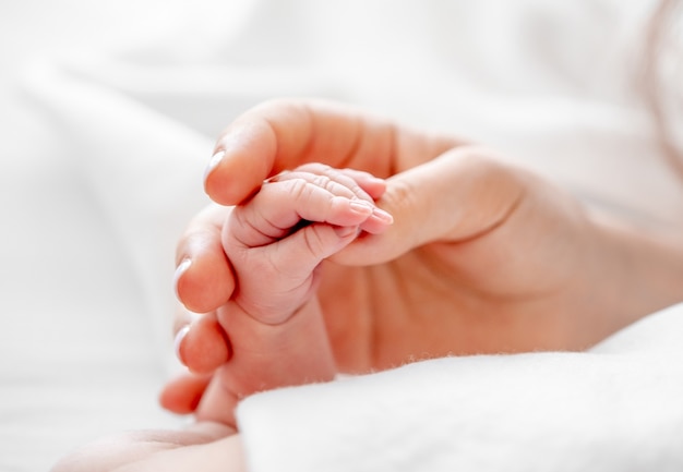 Photo mother holding tiny newborn baby hand with little fingers closeup in the bed with white bedding with daylight. concept of matherinity love, protection and tenderness