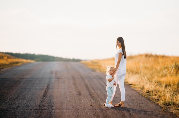 Mother holds her little daughter by the hand, they stand on the road in the rays of a summer sunset