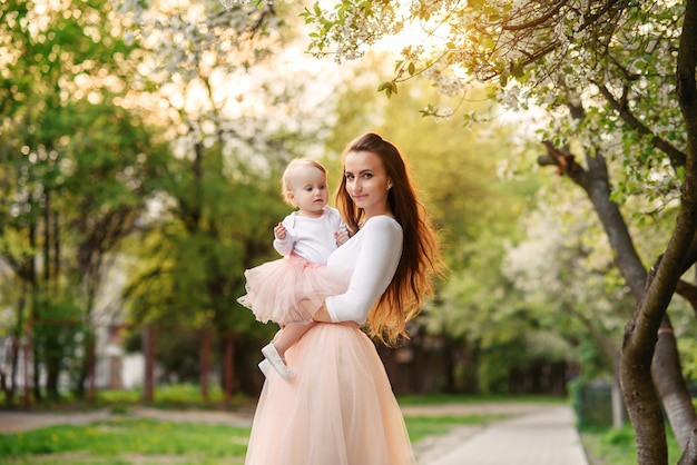 La madre tiene la sua piccola figlia tra le braccia tra alberi in fiore. mamma e il suo bambino indossavano un abito rosa dall'aspetto familiare.